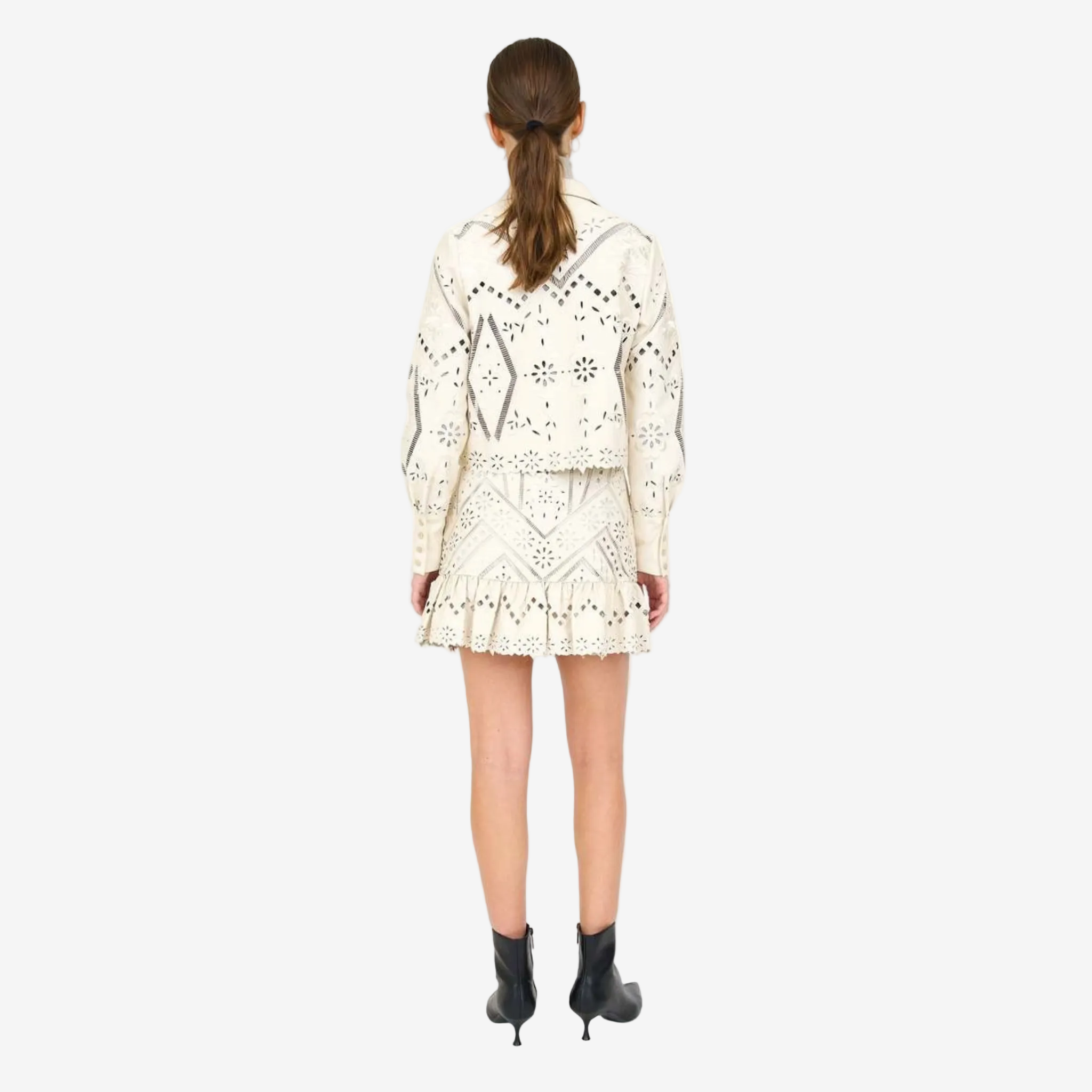Back view of a cream‑colored laser‑cut jacket with floral and geometric cutout patterns, paired with a matching mini skirt featuring a ruffled hem and worn with black ankle boots.