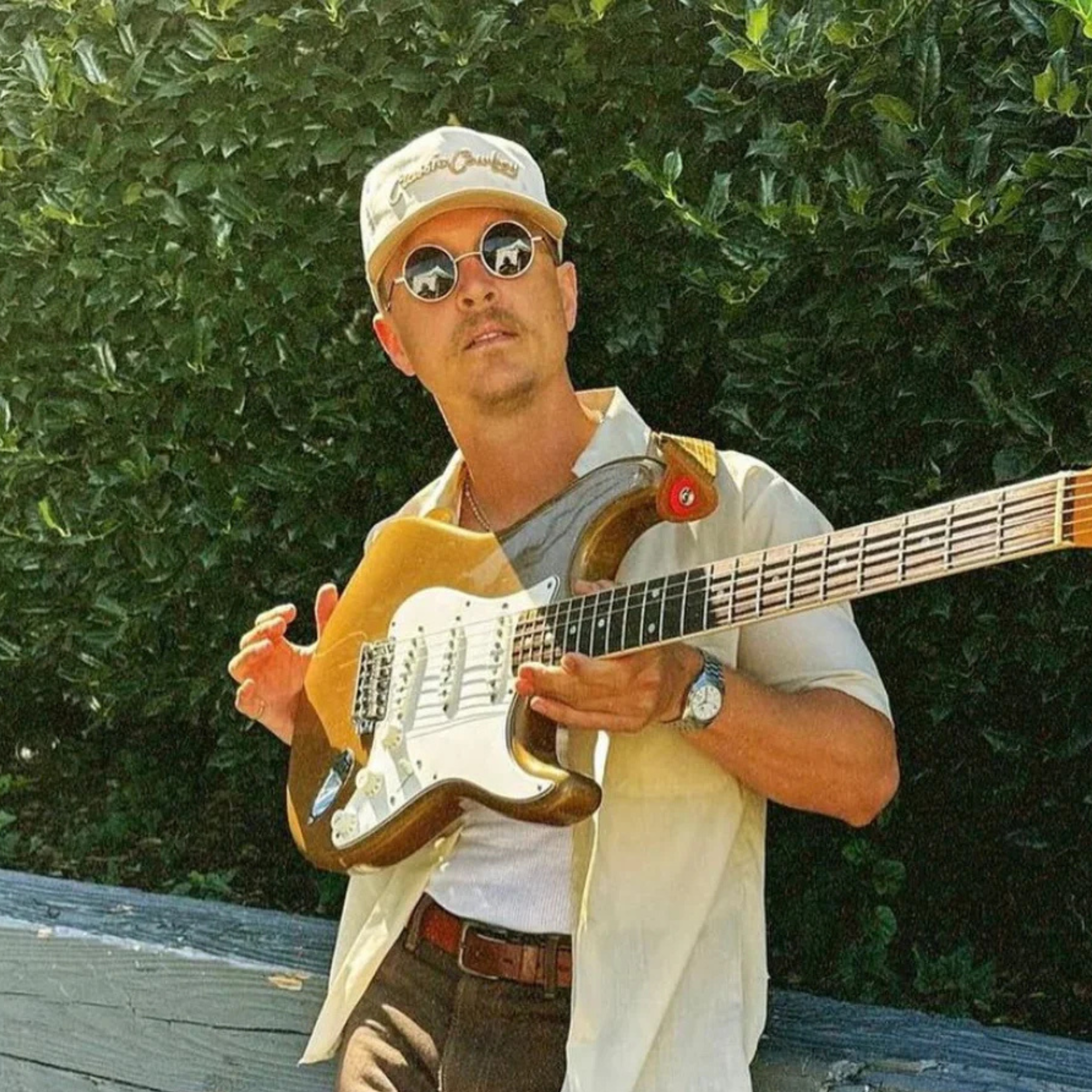Man playing a guitar outdoors with greenery in the background